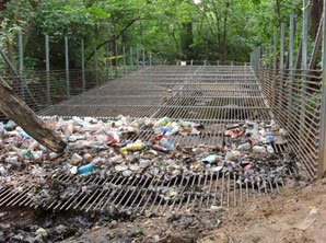 Large, metal cage-like structure with cage on bottom and two sides crossing a river and filled with trash